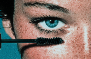 Close-up of a mascara wand being applied to an eye with a teal background