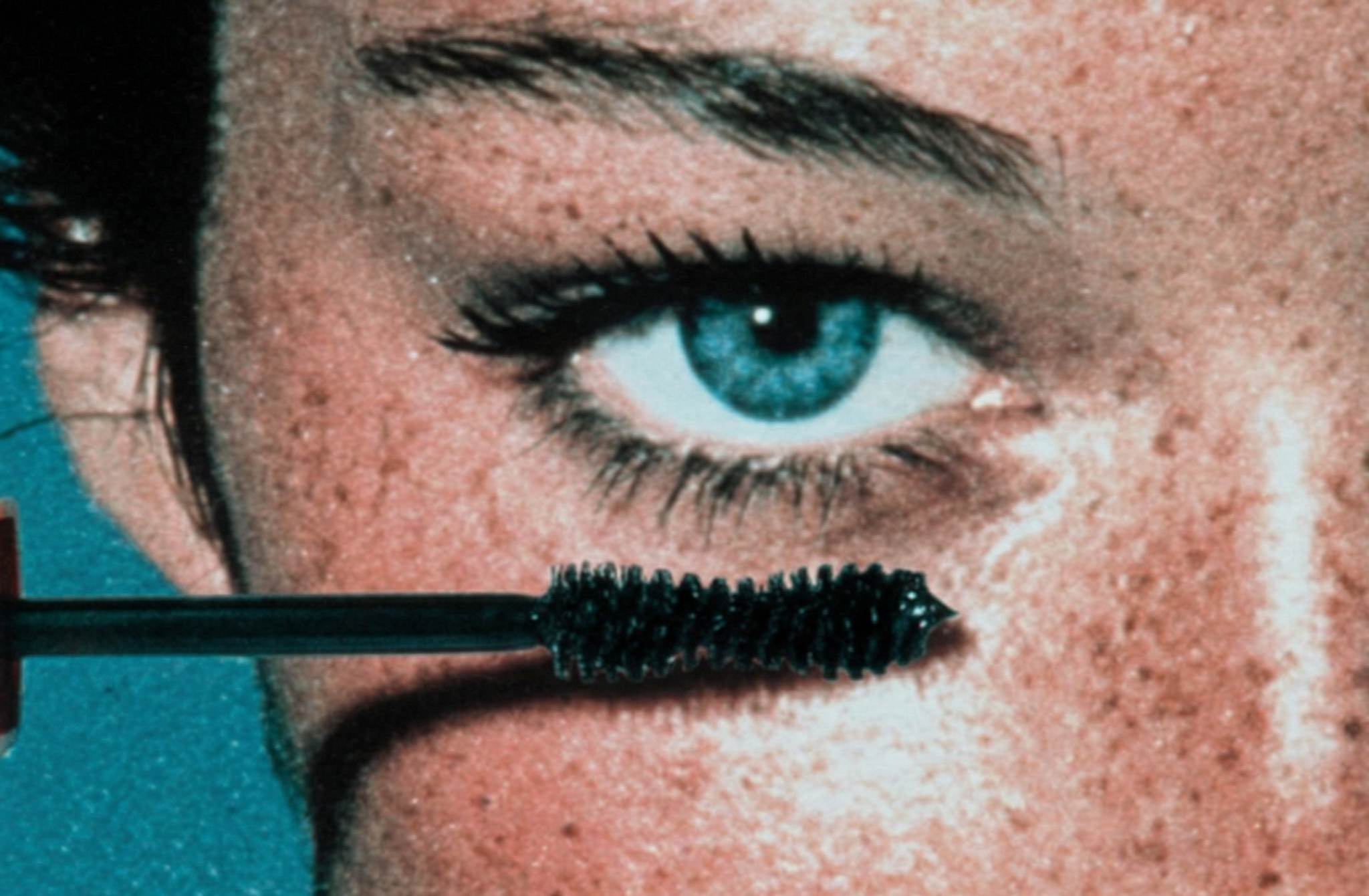 Close-up of a mascara wand being applied to an eye with a teal background